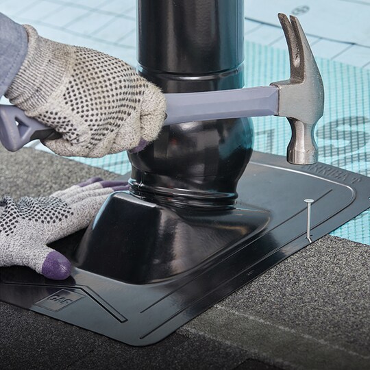 Shaded image of a GAF roofer’s gloved hands using a hammer and nail to install Master Flow pivot pipe boot flashing to a residential roof